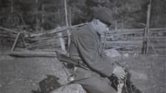 Mies (metsänhoitaja Torsten Rosenbröijer) istuu kivellä haulikko selässä ja rapsuttaa ajokoiraa.
