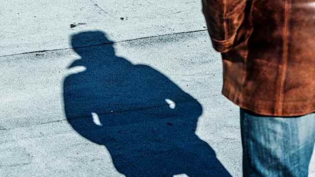 Seen from the side,  a man in jeans and a brown leather jacket. Most of the photo shows the shadow of his entire body on the pavement.