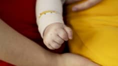 A baby's fist with parts of an adult arm and hand and red and yellow clothing around it.