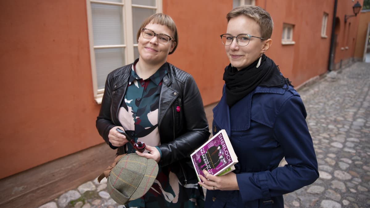 Tutkija Elina Karvo ja Suvi Laine poseeraavat Sherlock Holmesin hatun ja kirjojen kanssa Vanhan Suurtorin maisemassa.