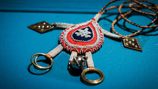 Käsintehty koru, jossa on punainen ja sininen koristeellinen keskiosa. Korun yläosassa on punottu nauha ja alaosassa metallisia yksityiskohtia.