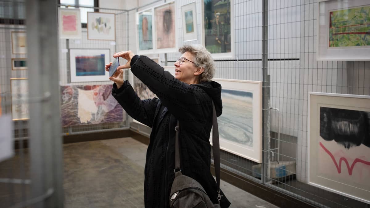 Nainen kuvaa puhelimella taidemaalauksia täynä olevassa tilassa.