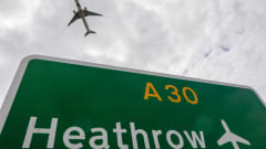 Kyltti, jossa lukee lentokentän nimi. Taivaalla lentävä lentokone.