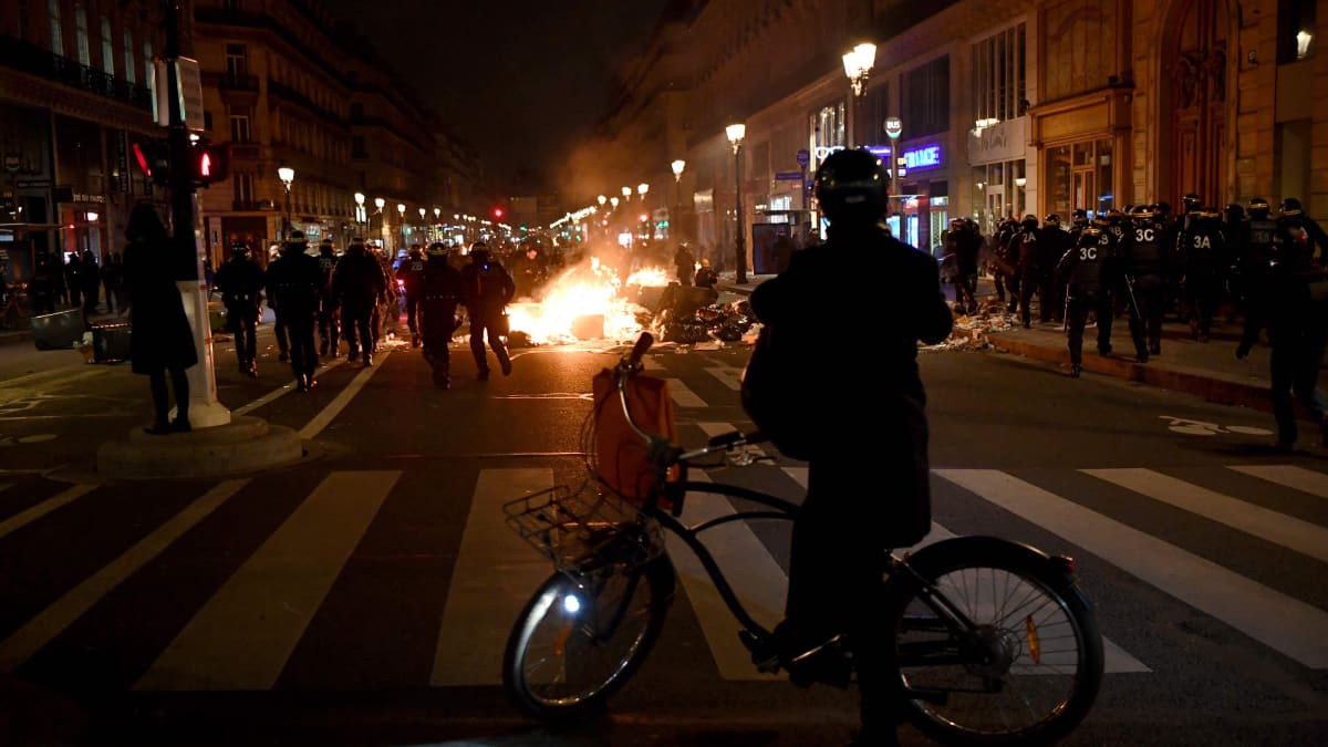 Pyöräilijä seisoo kuvan etuosassa katsomassa taustalla palavaa mielenosoittajien sytyttämää paloa.