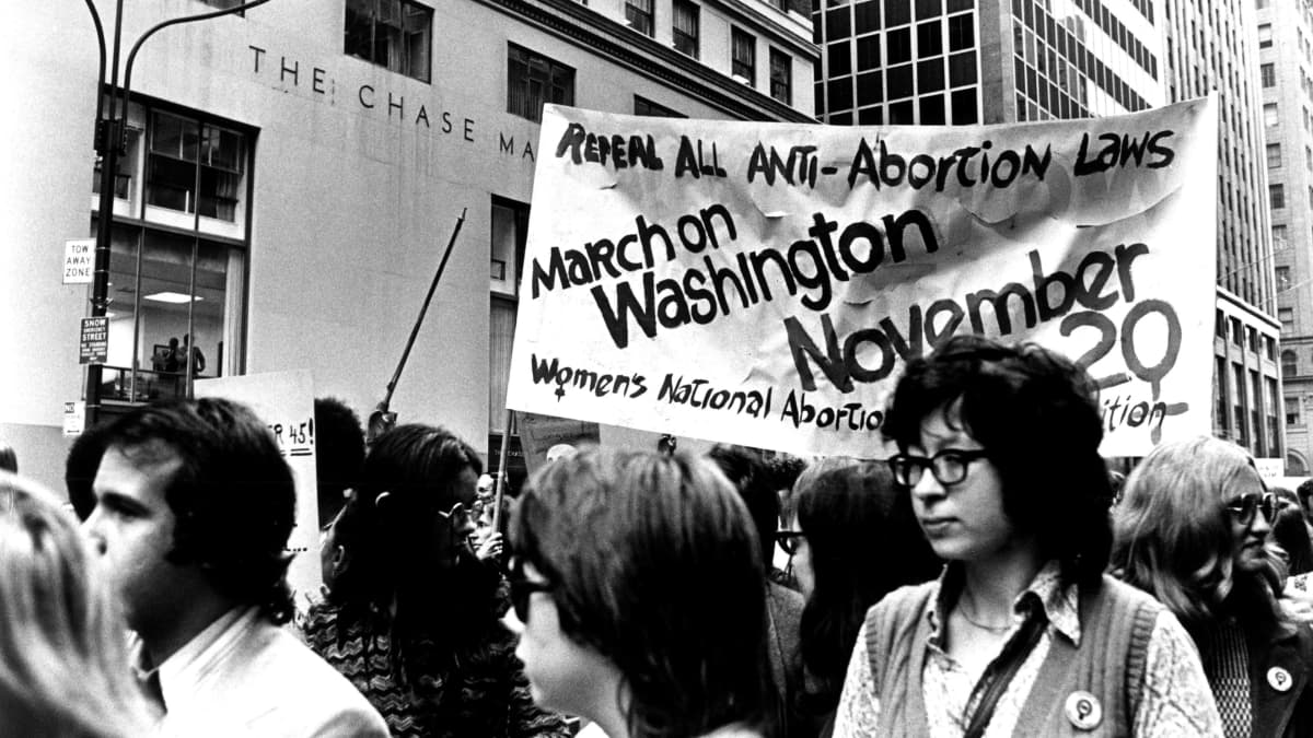 Mustavalkoisessa kuvassa nuoria naisia ja miehiä marssii aborttioikeutta vaativan banderollin kanssa. 