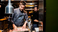 Two men wearing dark clothing in a restaurant kitchen preparing food.