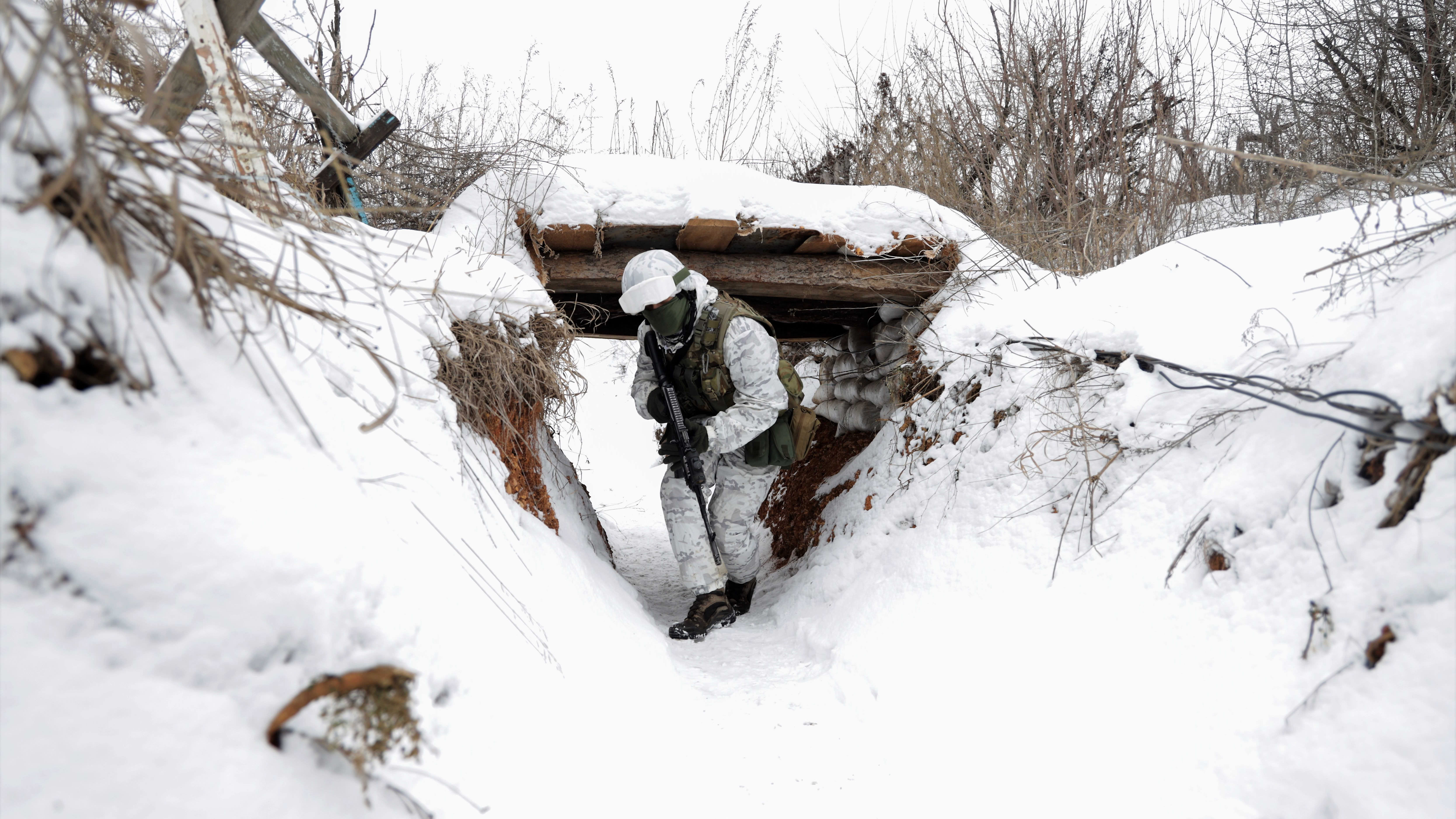Lumipukuun pukeutunut sotilas kulkee jonkinlaisen rynnäkkökiväärin kanssa lumisessa matalassa juoksuhaudassa. Lumen alta törröttää siellä täällä heiniä.