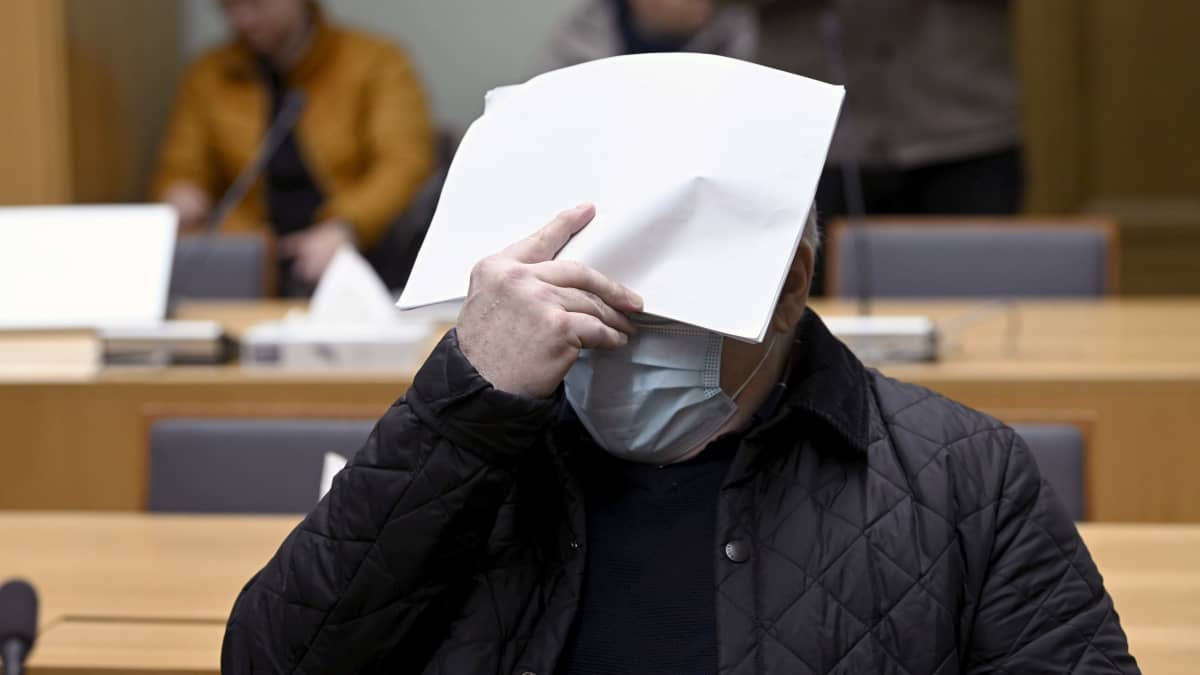 Kasvomaskiin pukeutunut henkilö peittää kasvonsa paperiarkilla oikeussalissa.