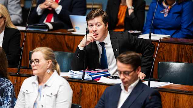 A man wearing a black blazer and blue tie sits in Parliament with other MPs seated around him.