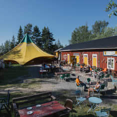 Ihmisiä istuskelee kesäpäivänä punaiseksi maalatun kahvilan terassilla. Taaempana näkyy keltainen sirkusteltta, vastapäätä keltainen omakotitalo.