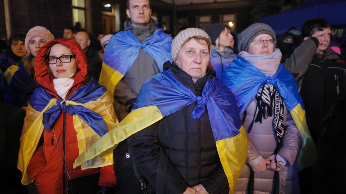 Vakavailmeisiä ihmisiä mielenosoituksessa, monilla pipo päässä ja Ukrainan lippu hartijoiden ympärillä. 