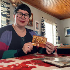 Kati Rita selailee vanhoja kuvia pirtin pöydän ääressä Ristijärvellä ja esittelee yhtä niistä kameralle.