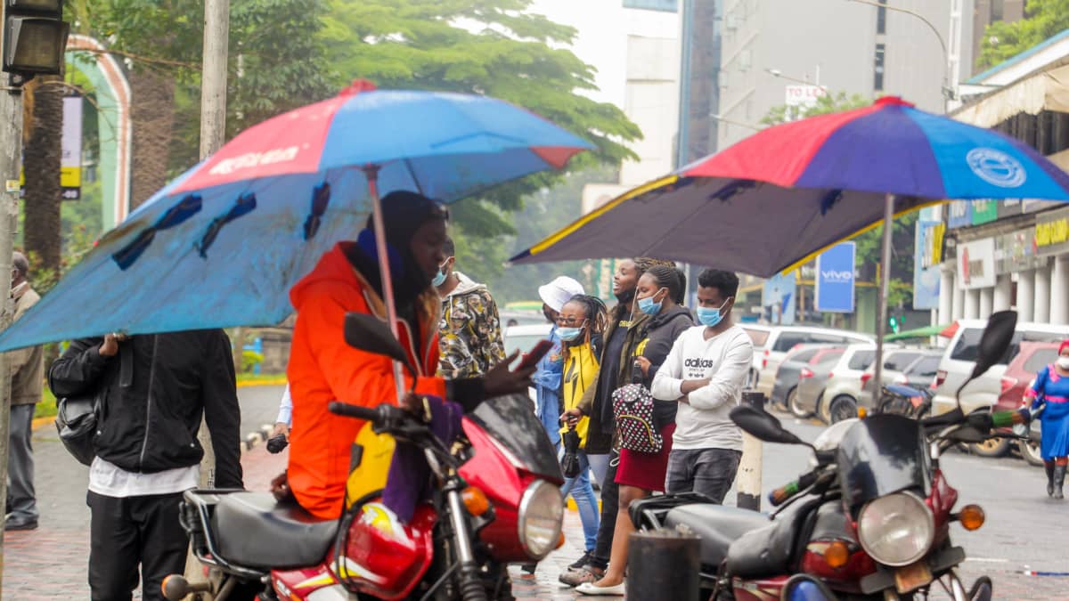 Ihmisiä seisoo kadulla kirurgiset kasvomaskit kasvoilla. Etualalla Kaksi moottoripyörää päivänvarjojen alla.