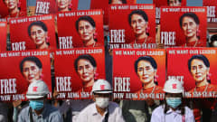 Mielenosoittajia kypärät päässä, taustalla seinänä punaisia kylttejä, joissa Aung San Suu Kyin kuva ja vaatimus hänen vapauttamisestaan.