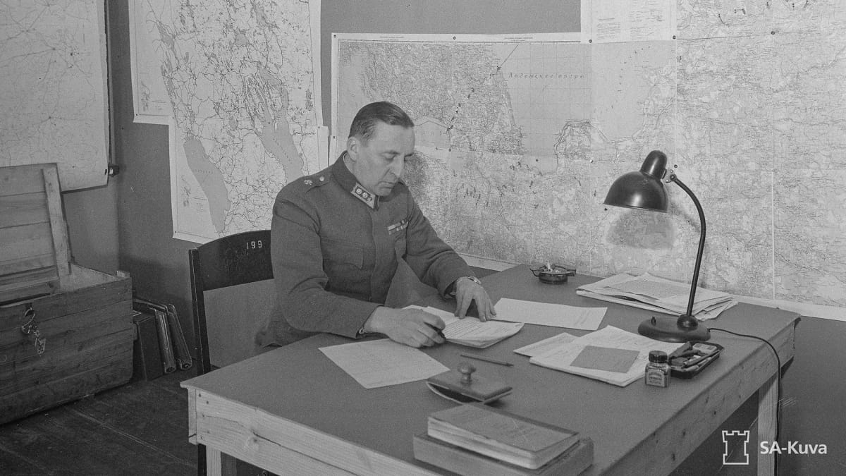 Reino Hallamaa tutkiskelee papereita työpöydän äärellä vanhassa sota-ajan valokuvassa.