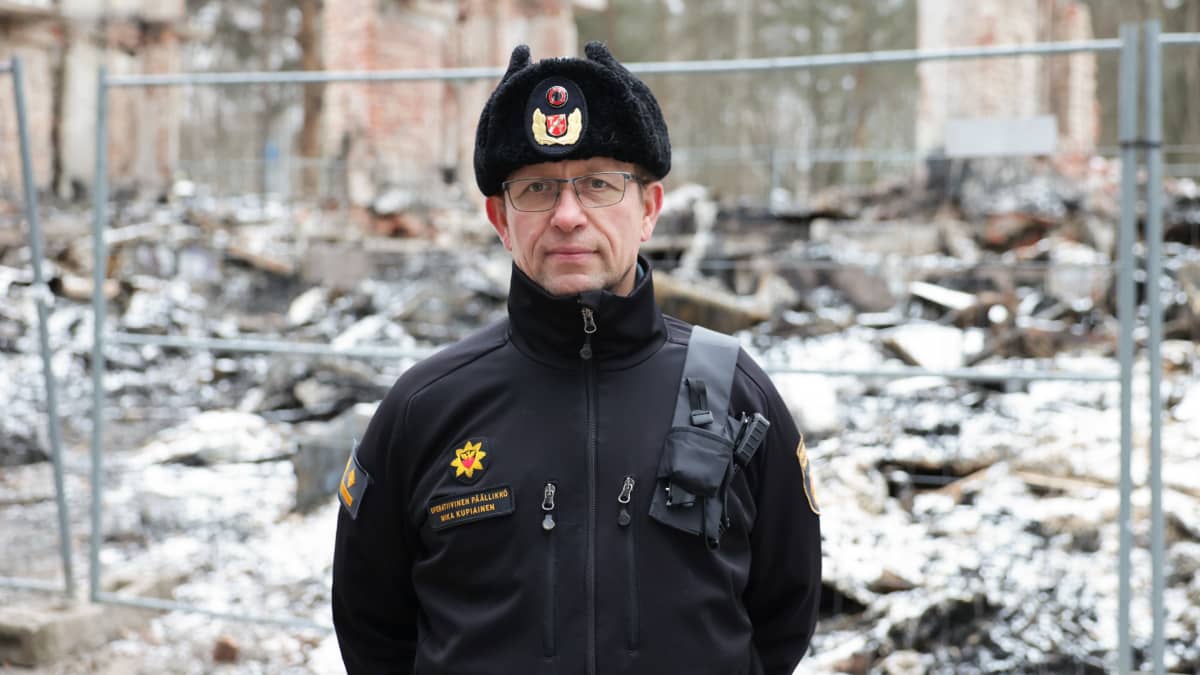Pirkanmaan pelastuslaitoksen operatiivinen päällikkö Mika Kupiainen seisoo virkapuvussa Hipposkylän poltetun talon raunioiden edessä, hymyilee kameralle.