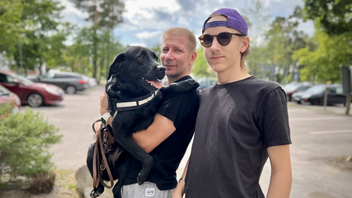 Musta labradorinnoutaja omistajansa Janne Kossilan sylissä, vieressä Jannen poika Nico Kossila aurinkolasit päässä ja lippis väärinpäin.