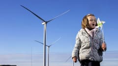 Kolme suurta tuulimyllyä seisoo maisemassa sinistä taivasta vasten. Etualalla pikkutyttö puhaltaa kädessään olevaan tuulihyrrään.