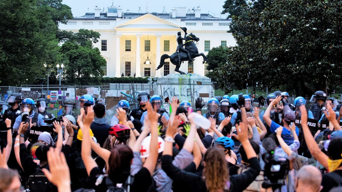 Mielenosoittajien joukko seisoo mellakkavarusteissa olevan poliisiketjun edessä ja pitää käsiään ylhäällä. Taustalla näkyy Valkoinen talo ja Andrew Jacksonin patsas.