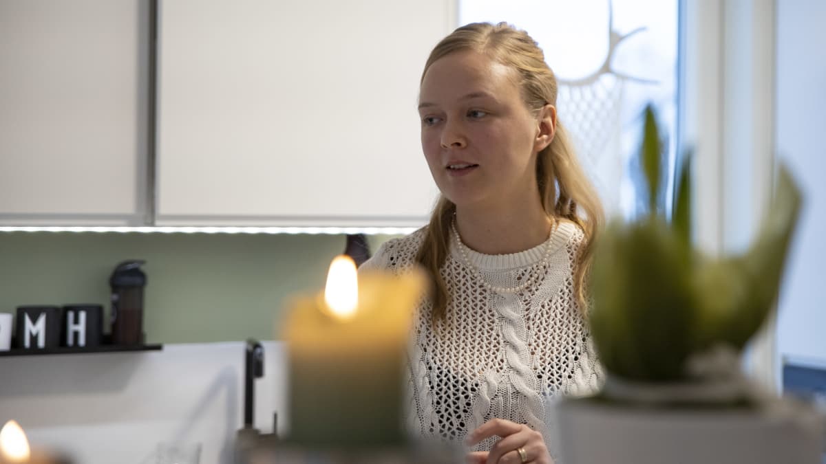 Sijaisvanhempi Mari Kortesoja istuu keittiössä vaaleassa neuleessa, etualalla pöydällä palaa kynttilä.