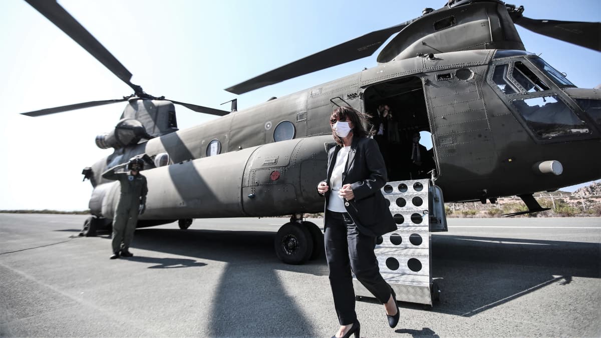 Kreikan presidentti Katerína Sakellaropoúlou on tulossa helikopterista. Hänellä on harmaa asu. Taustalla lentäjänasuinen kypäräpäinen mies vetää kättä lippaan presidentille.