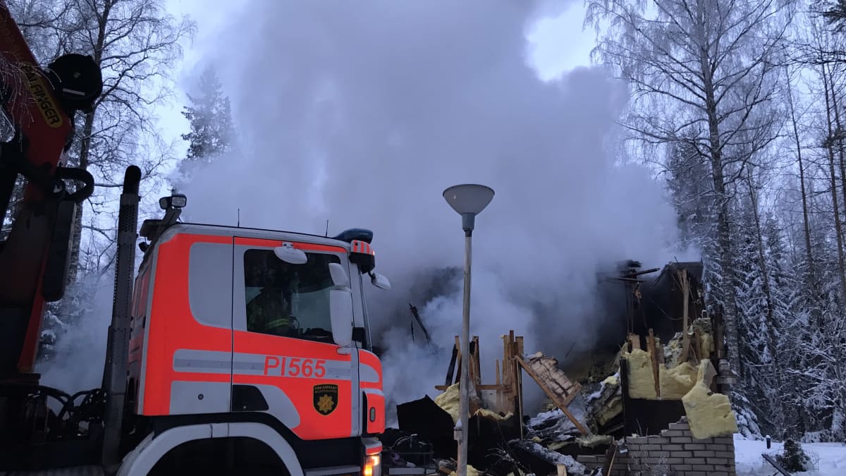 Paloauto tulipalossa tuhoutuneen talon edustalla. Raunioista nousee savuta.