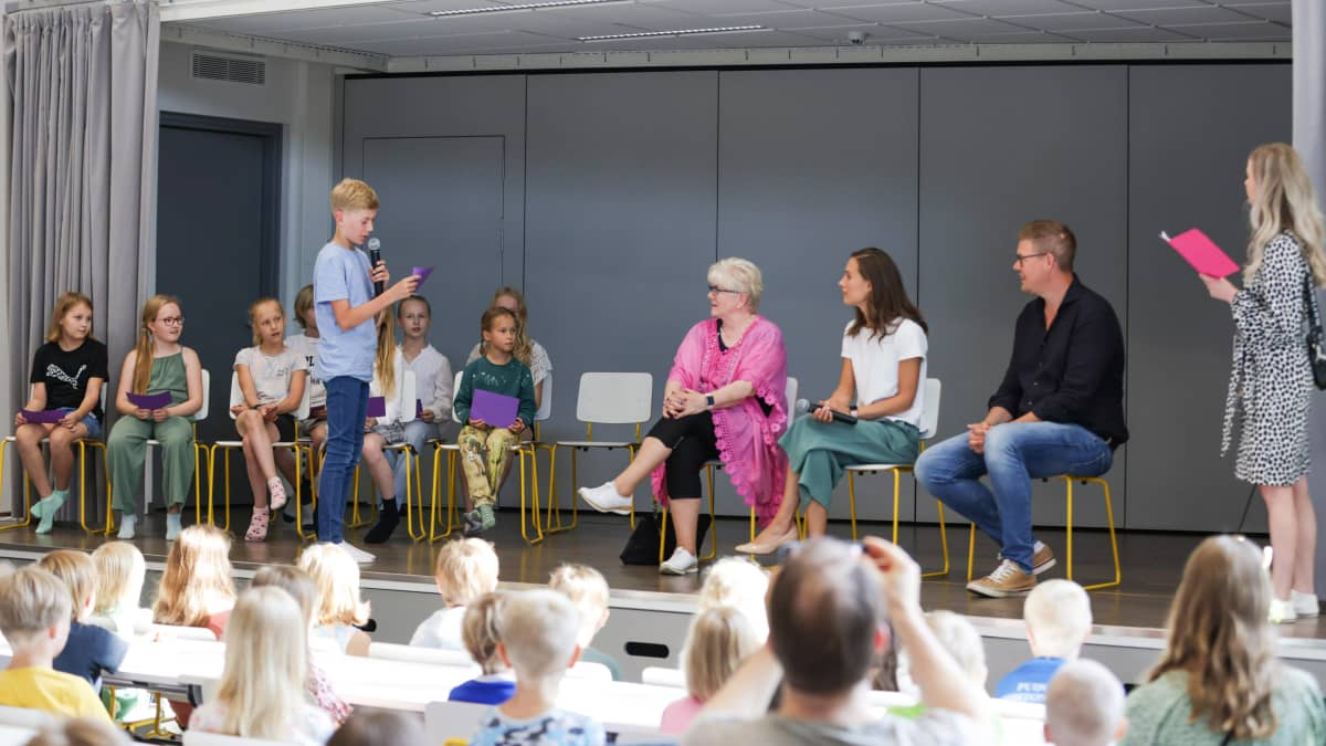 Kansanedsustaja Tuula Väätäinen, pääministeri Sanna Marin ja SDP:n eduskuntaryhmän puheenjohtaja Antti Lindtman istuvat lavalla alakoulun oppilaiden kanssa. Yksi lapsista kysyy kysymystä.