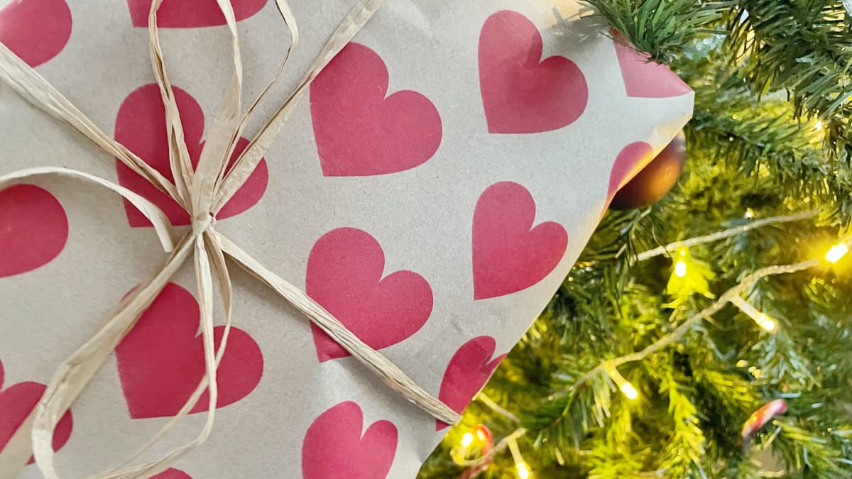Ett paket packat i vitt papper med rosa hjärtan på lutar mot en julgran med julljus.