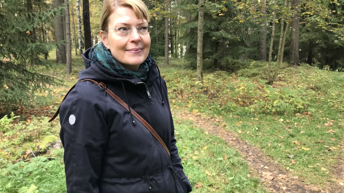 Pirkanmaan maakuntamuseon tutkija Kirsi Luoto seisoo metsässä Tampereen Kalevanharjulla. Luodolla on tummansininen anorakki ja turkoosi huivi.  