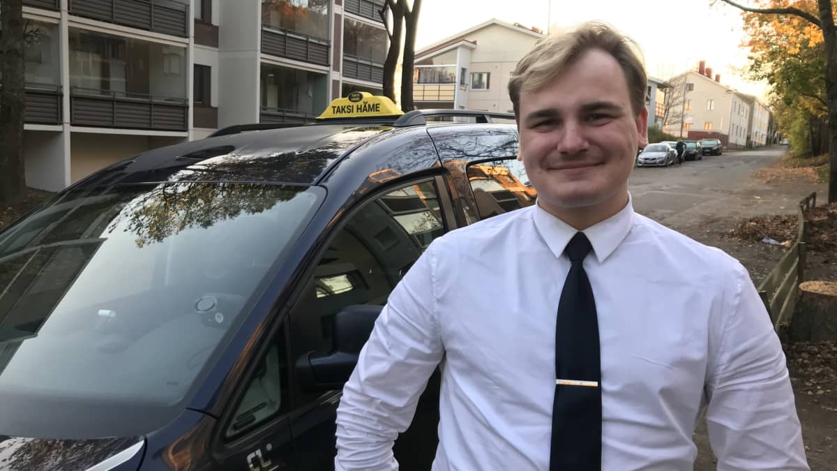 Hymyilevä taksinkuljetta seisoo autonsa edessä 