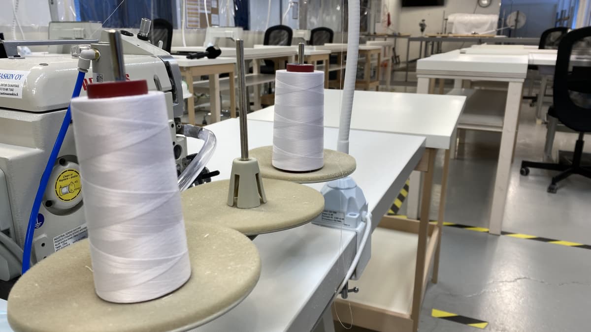 Teollisuushallissa pöydillä on ompelukoneita, lankaa ja kangasta suojavarusteiden ompelemista varten. 
