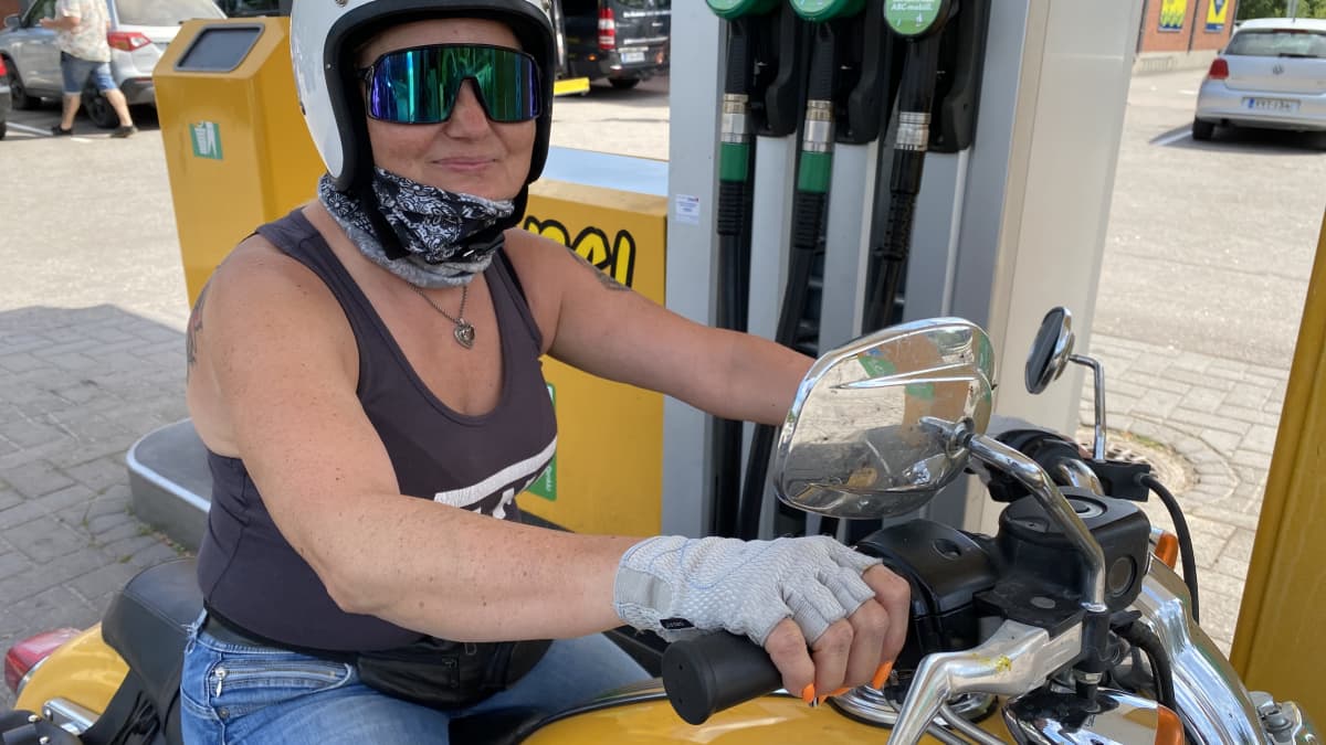 Nainen istuu keltaisen moottoripyörän selässä huoltoaseman polttoainepisteellä 