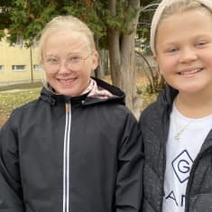 Seminaarin koulun viidesluokkalaiset, vaaleatukkaiset Kukka Oksanen ja Fia Hirvonen katsovat kameraan hymyillen syksyisellä koulun pihalla mustissa takeissaan. Kuva otettu 11.10.2021.