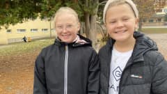 Seminaarin koulun viidesluokkalaiset, vaaleatukkaiset Kukka Oksanen ja Fia Hirvonen katsovat kameraan hymyillen syksyisellä koulun pihalla mustissa takeissaan. Kuva otettu 11.10.2021.