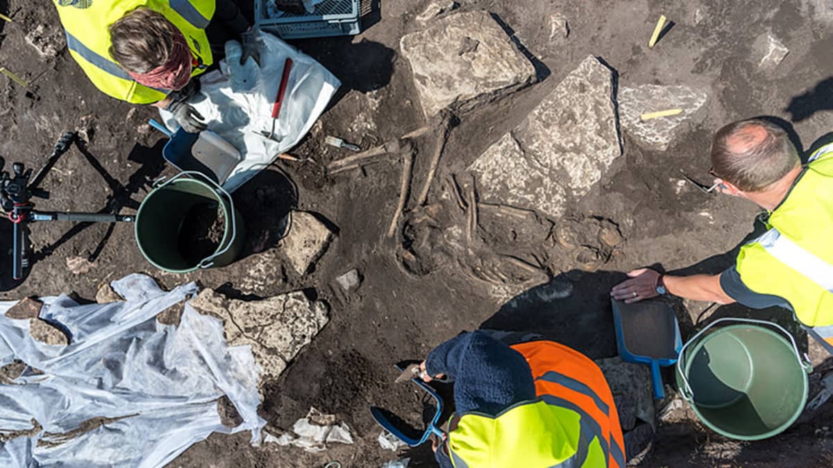 Ylhäältäpäin otettu kuva kolmesta arkeologista kavamassa esiin kivien välissä sikiöasennossa makaavaa luurankoa. esta