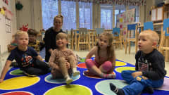 Neljä lasta istuu lattialla päiväkodissa. Taustalla lastenhoitaja.