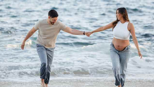 Ett par promenerar hand i hand längs strandkanten, med havet i bakgrunden. Kvinnan bär en vit topp som visar hennes gravidmage.