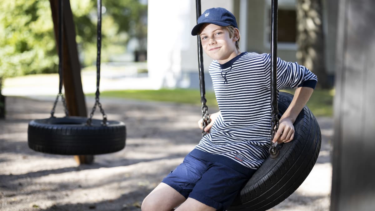 Poika istuu rengaskeinussa aurinkoisena päivänä sininen lippis päässään ja hymyilee.