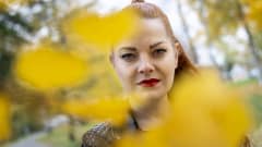 Nainen katsoo suoraan kameraan syksyisten lehtien takaa