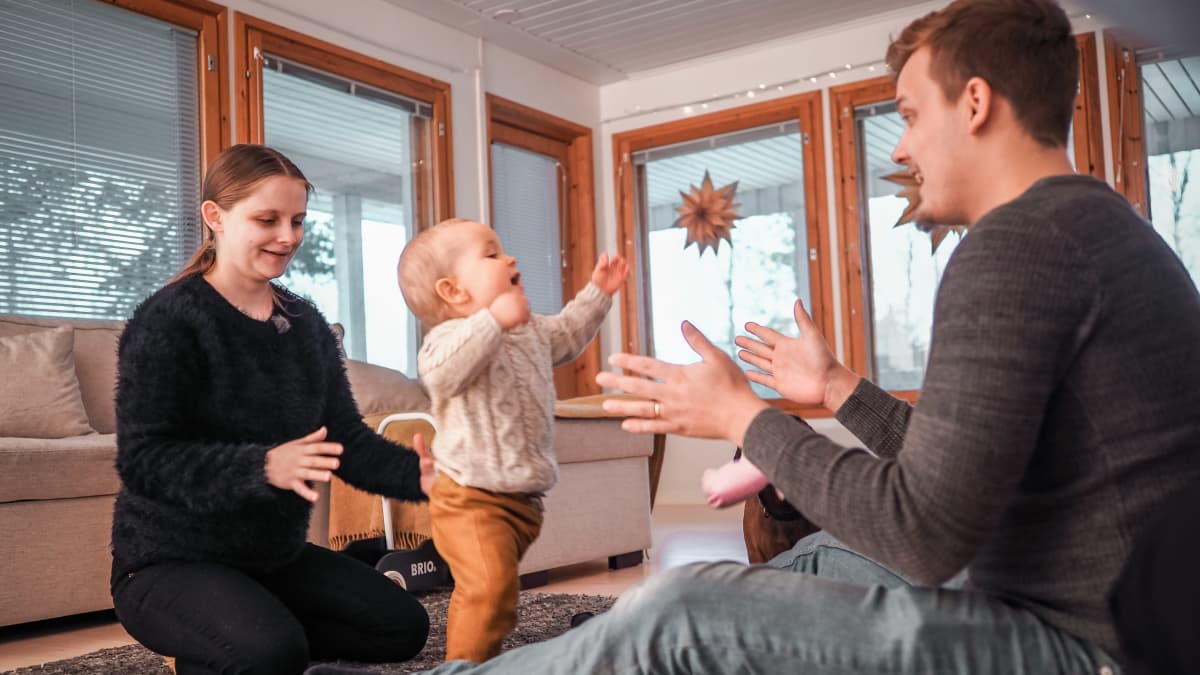 Vanhemmat ja juuri kävelemään oppinut lapsi leikkivät olohuoneen lattialla.