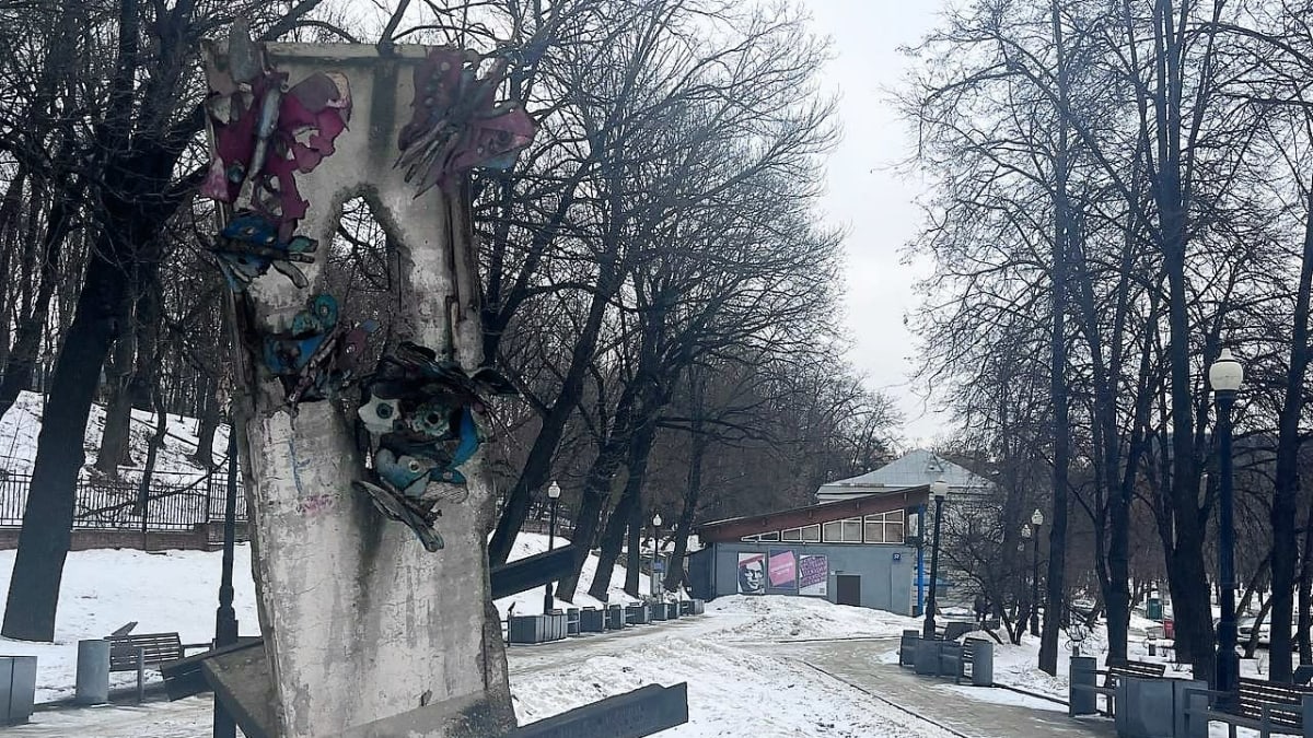 Rikkonainen betonilaatta on pystytetty hieman vinosti metalliselle jalustalle puistoon. Kauempana näkyy rakennus, jonka seinässä on Andrei Saharovin kuva. Maisema on luminen mutta ruohoa pilkottaa lumen alta.