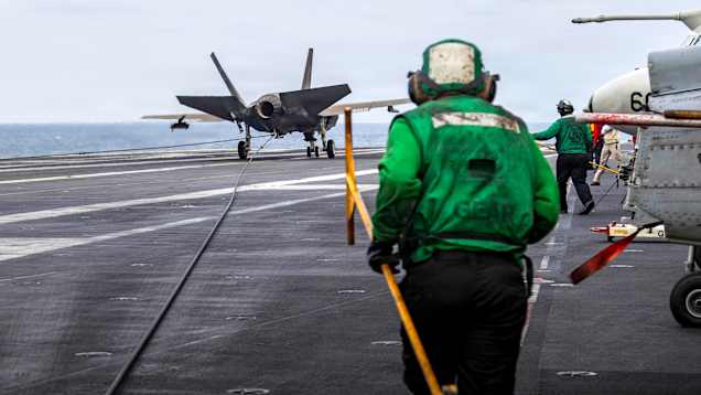 Hävittäjä lentotukialuksen kannella. Etualalla seisoo henkilö kuulokkeet korvillaan.