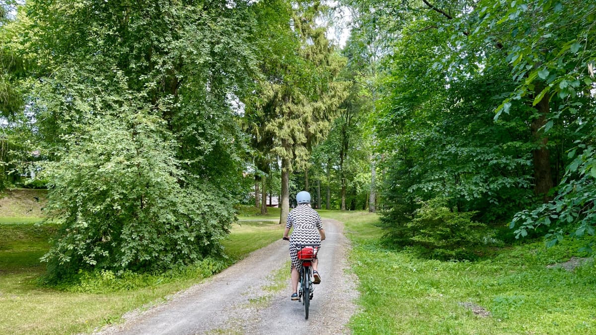 Mustavalkoiseen kesämekkoon pukeutunut nainen ajaa polkupyörällä poispäin vihreällä puistotiellä