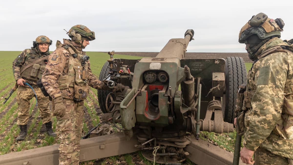 Kolme ukranailaissotilasta maastopuvuissa ja kypärissä valmistautuu ampumaan tykillä, vihreää peltoa. 