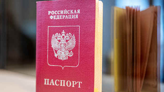 A red Russian passport with Cyrillic lettering standing on a shelf, with a mirror image of the document's open pages seen in the background.