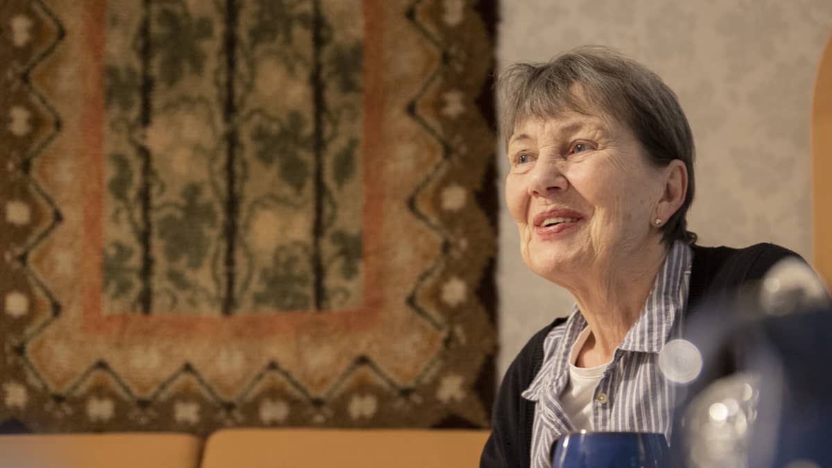 80 vuotta täyttävä Riitta Paterson istuu kahvipöydässä ja hymyilee. Taustalla karjalaista perintöä oleva ryijy.