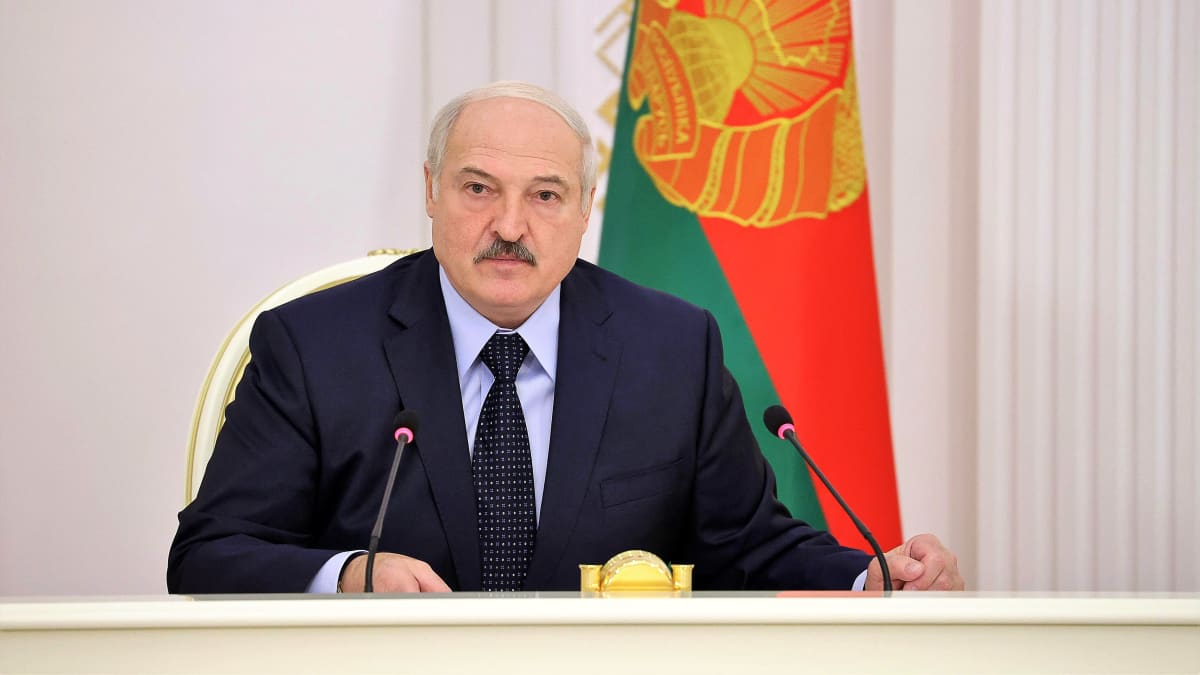 Lukashenka istuu pöydän takana tummassa puvussa ja kravatissa. Seinä on valkoinen, taustalla näky myös 