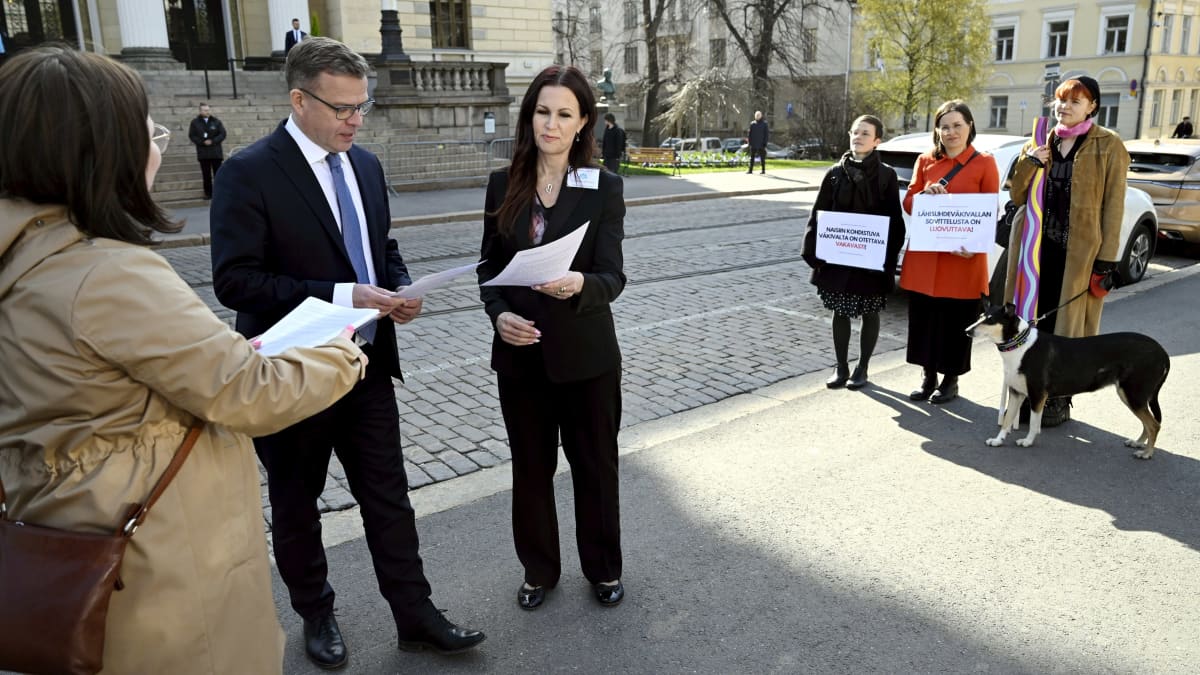 Petteri Orpo ja Pihla Keto-Huovinen tutkivat vetoomusta. Taustalla on Säätytalo ja kolme mielenosoittajaa kyltteineen.