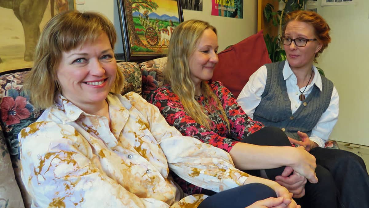 Kolme hymyilevää tutkijaa istuu kukikkaalla sohvalla työhuoneessa, jonka seinällä on värikkäitä tauluja ja julisteita. 