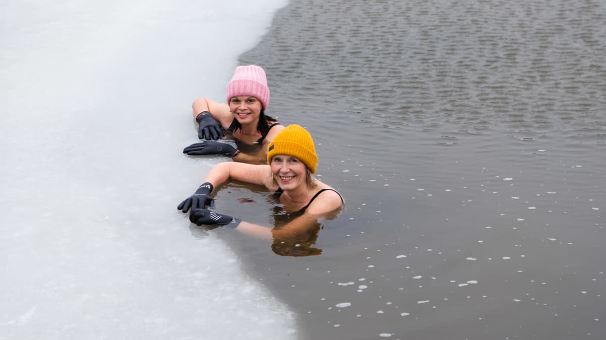 Riikka Airo ja Minna Lehtonen nojaavat suuressa avannossa jään reunaan. Päässä on pipot ja kädessä hanskat.